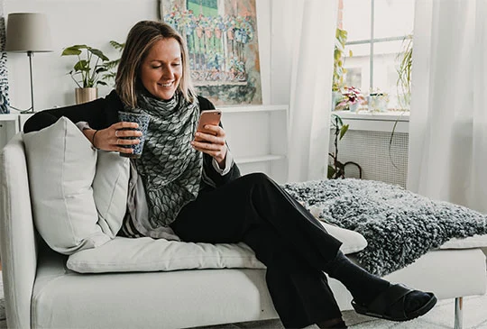 Kvinna dricker kaffe i soffan och tittar på mobilen